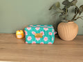 A green floral makeup bag with a gold label sits on a wooden surface next to a beige vase with green leaves and a lit candle.