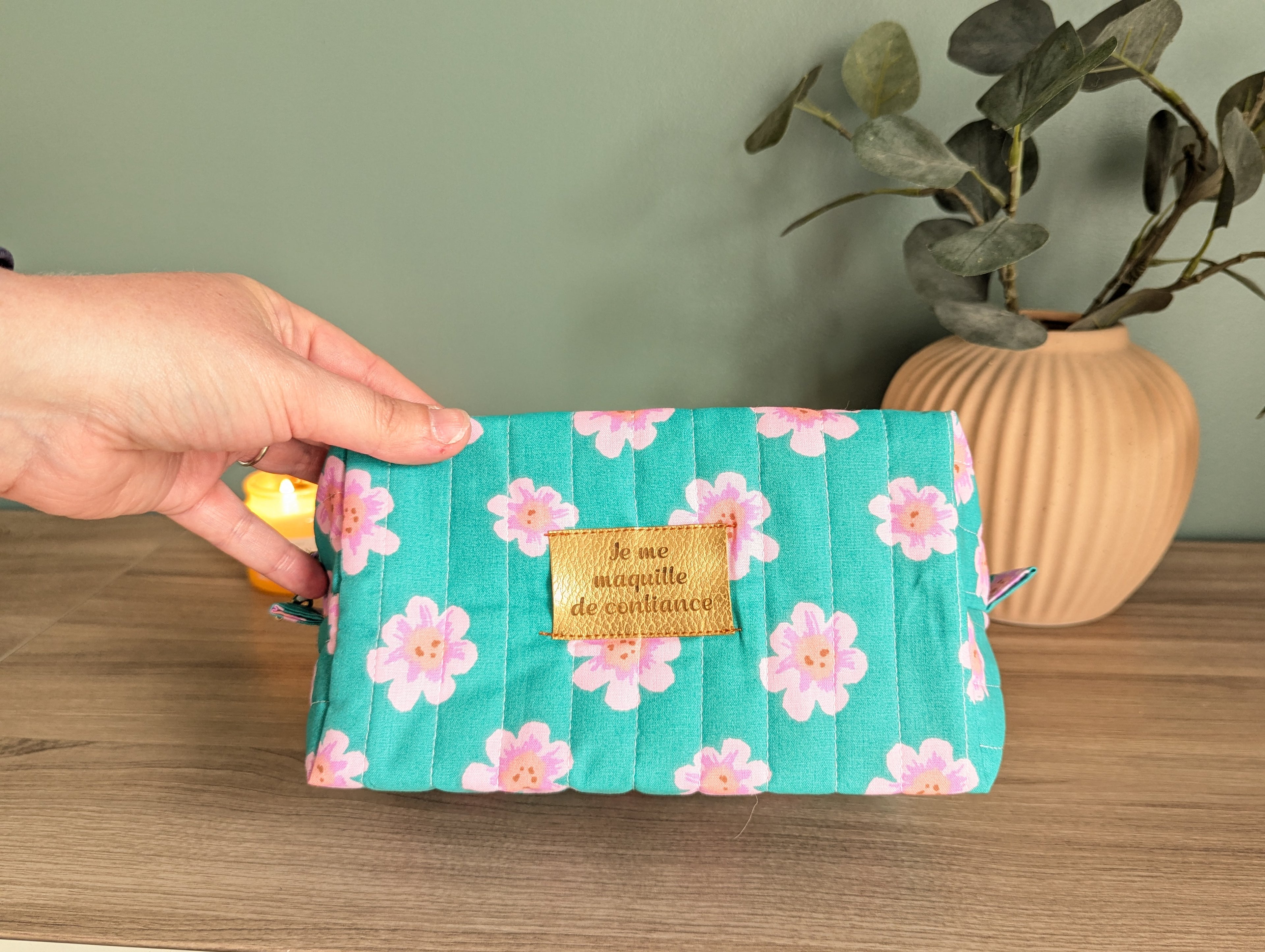 A hand holding a green makeup bag with a floral pattern, placed on a wooden surface next to a potted plant.