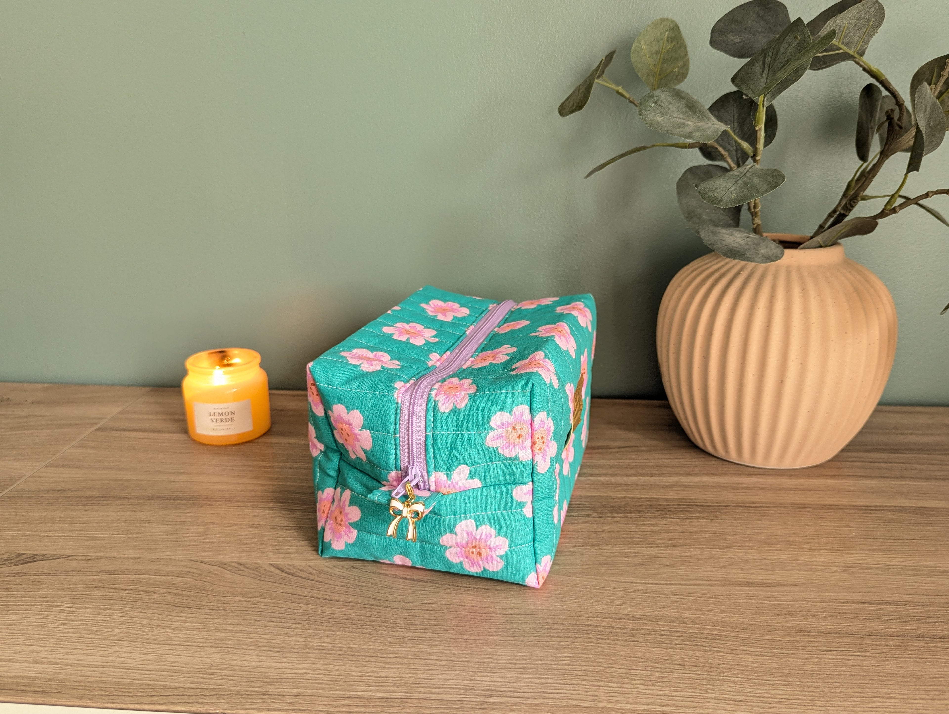 A small, square-shaped floral-patterned makeup bag sits on a wooden surface next to a small vase with green leaves.