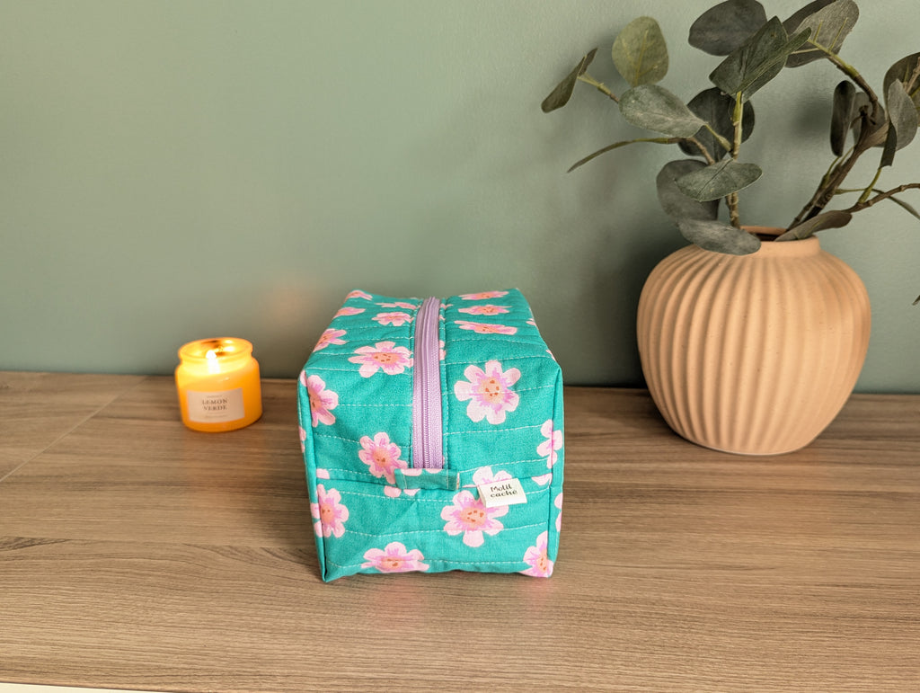 A teal floral makeup bag with a purple zipper sits on a wooden surface, accompanied by a small yellow candle and a potted plant.