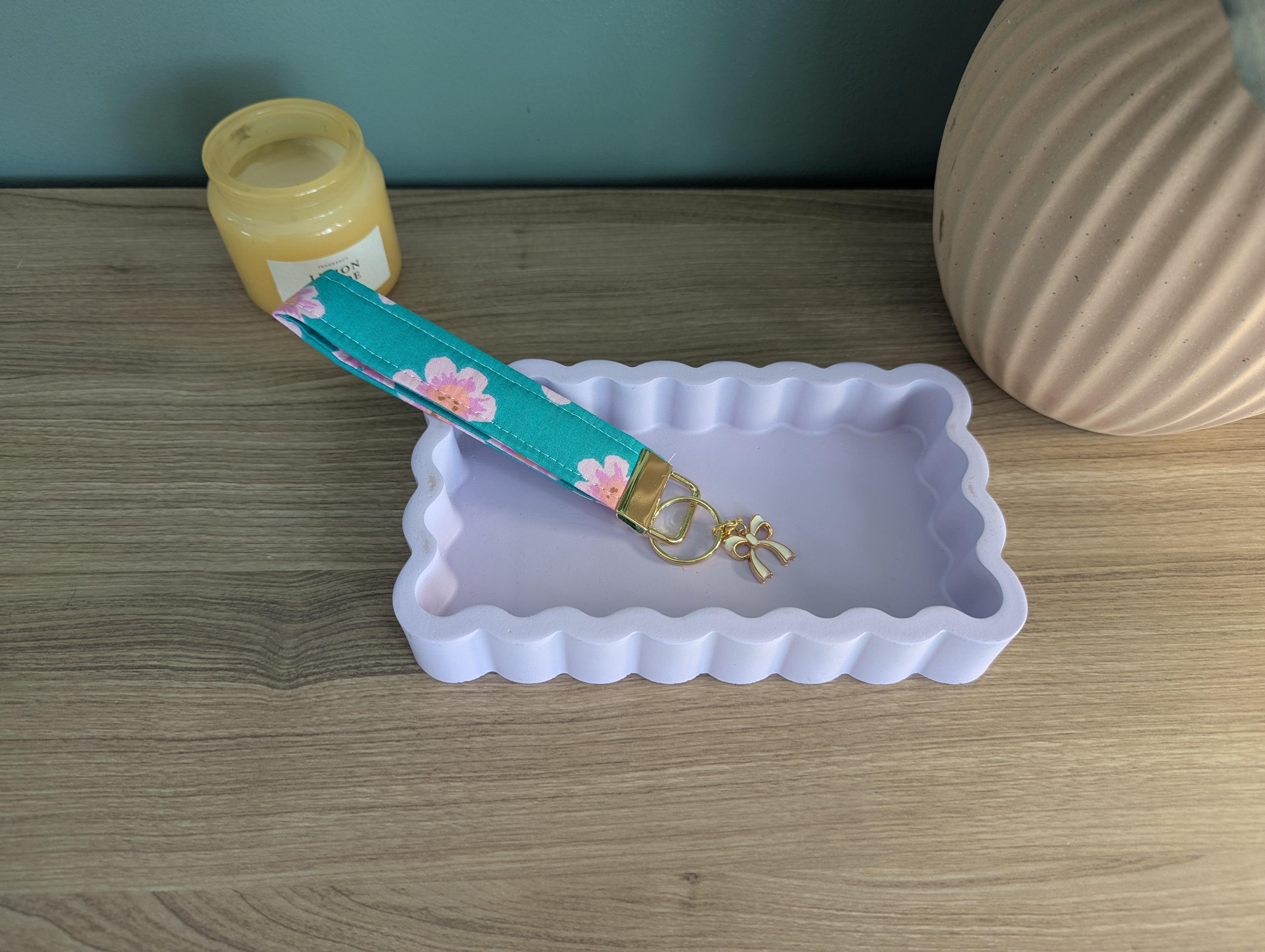 a blue and pink floral keychain resting on a white, scalloped tray, with a small vase and a candle on a wooden surface in the background.