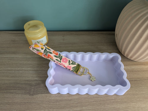 a pink and green floral keychain resting on a white rectangular tray, with a small white flower charm hanging from it.
