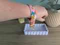 A person's hand is holding a colorful floral-patterned bracelet on a wooden surface, with a small white tray and a green plant in the background.