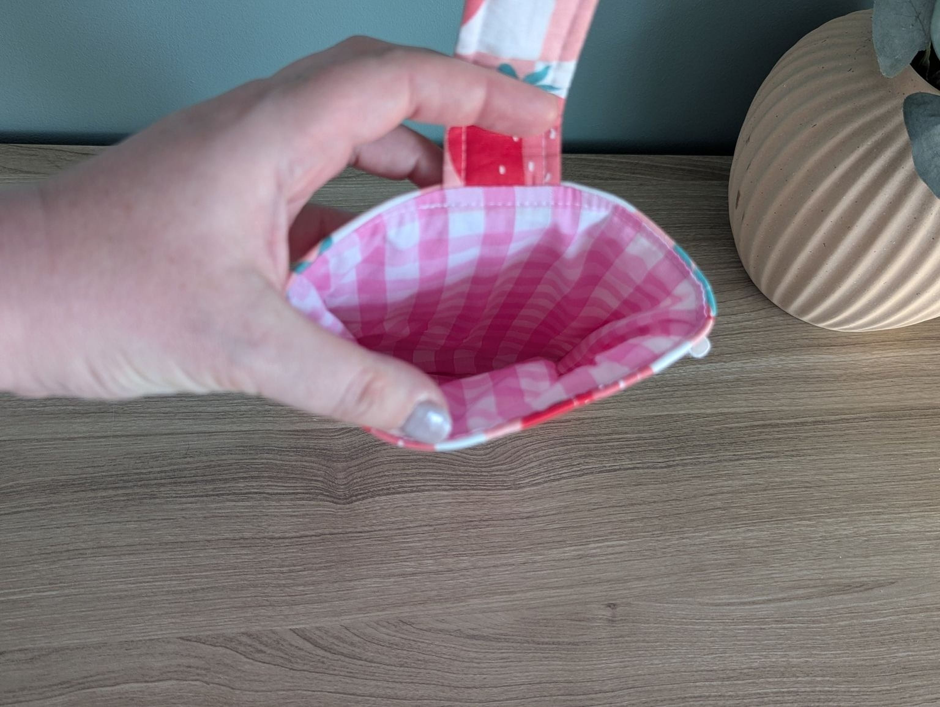 A hand holding a pink and white checkered pouch with a floral pattern, placed on a wooden surface next to a vase.