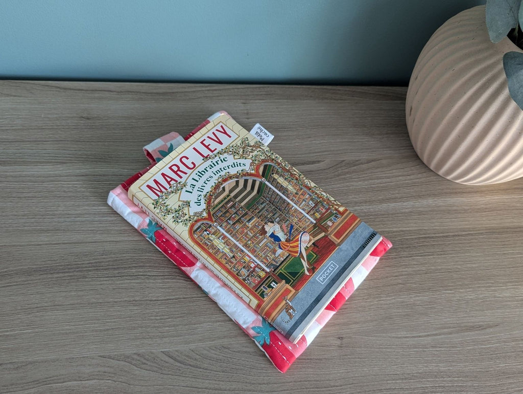a book titled "MARC LEVY" resting on a wooden surface, with a decorative book cover featuring a library scene. There is also a vase and a pink cloth visible in the background.