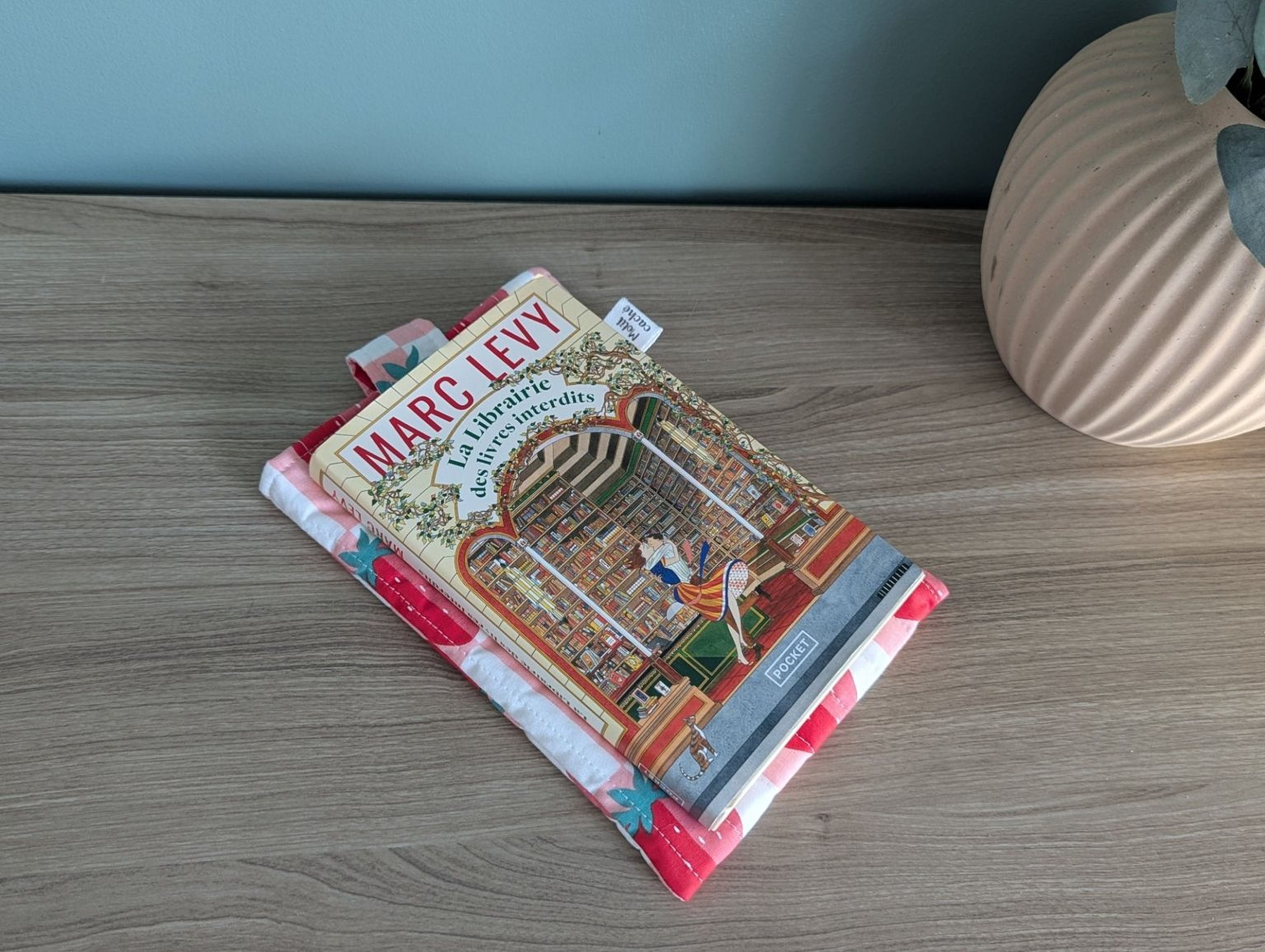 a book titled "MARC LEVY" resting on a wooden surface, with a decorative book cover featuring a library scene. There is also a vase and a pink cloth visible in the background.