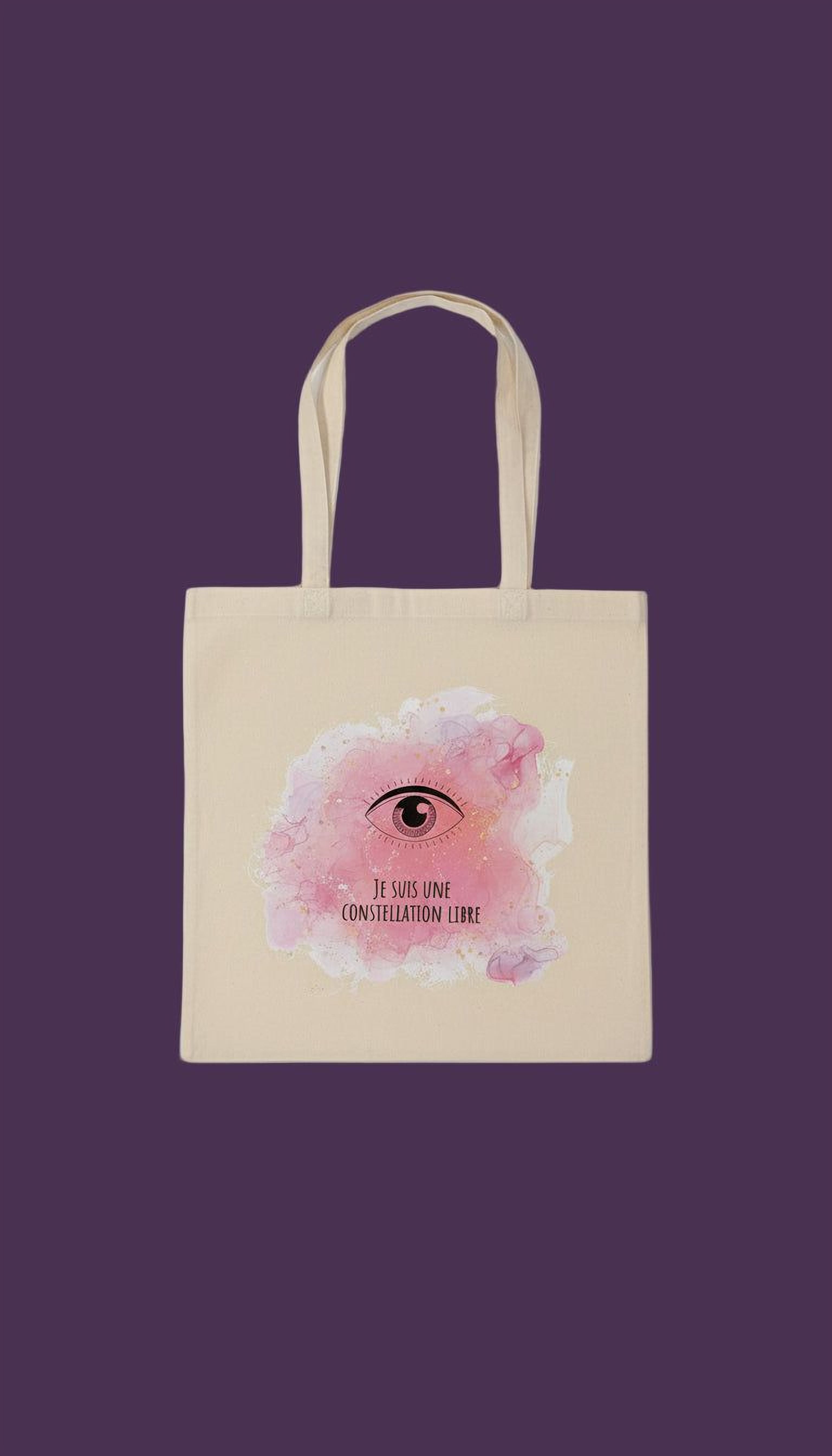 A beige tote bag with a pink and white watercolor design featuring an eye and the text "Je suis lune constellatione libre" (I am the constellation free).