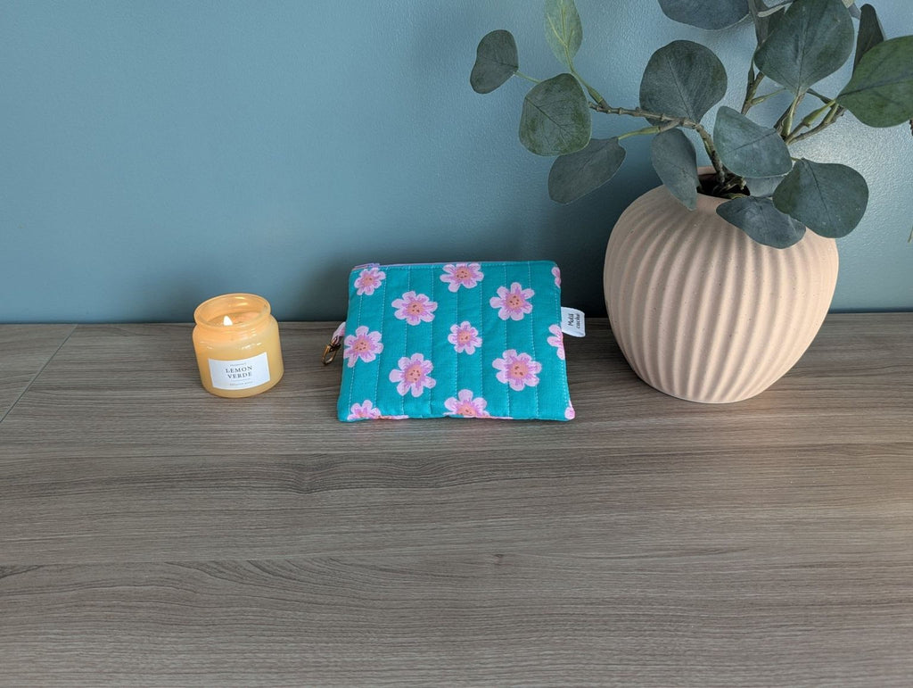 A blue floral patterned pouch, a yellow candle, and a white vase with green leaves are arranged on a wooden surface.