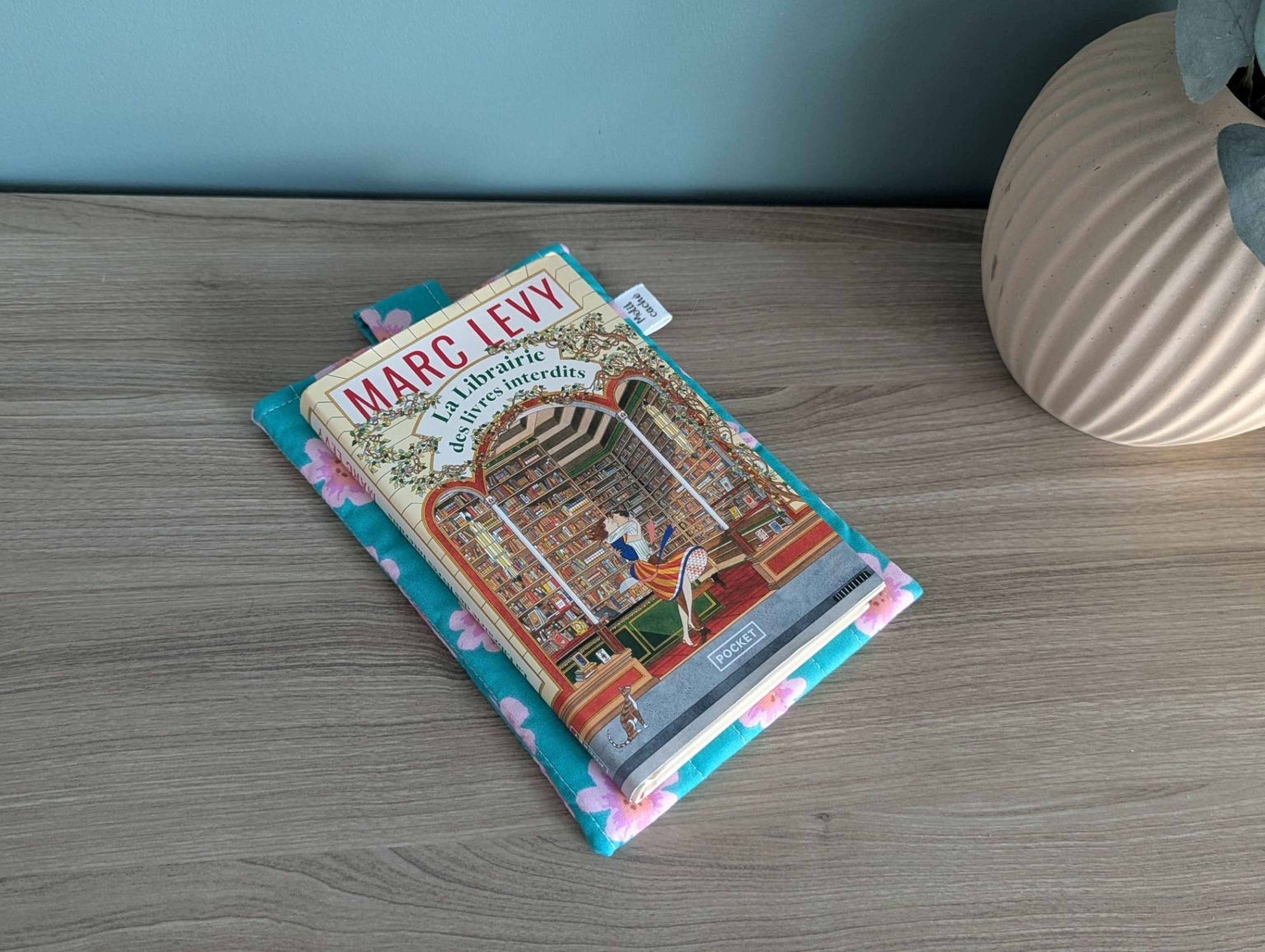 a book titled "MARG LEVY" resting on a blue and pink floral book cover, which is placed on a wooden surface. There is also a small white object, possibly a bookmark, next to the book.