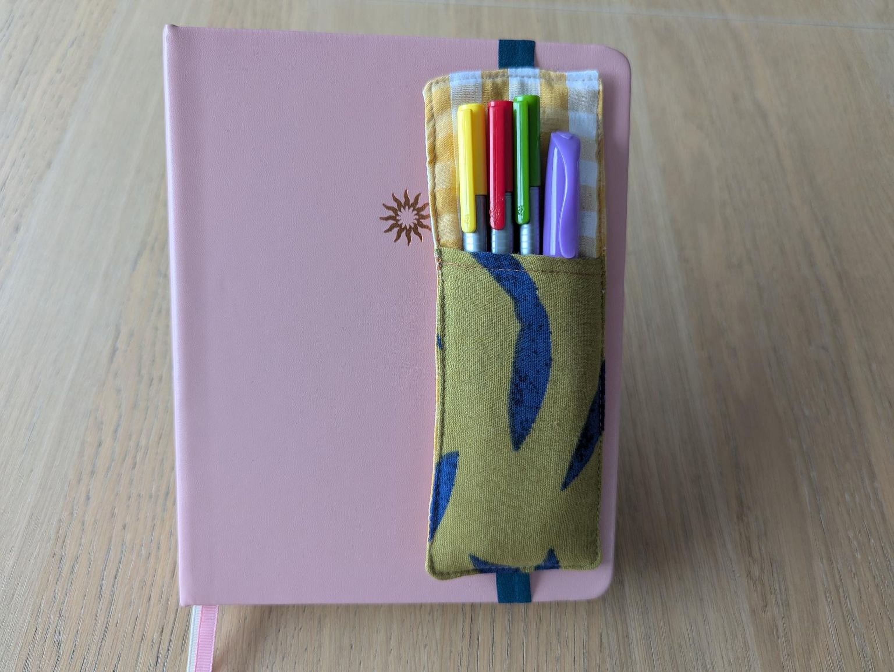 A pink notebook with a pocket attached to it, containing a variety of colorful pens.