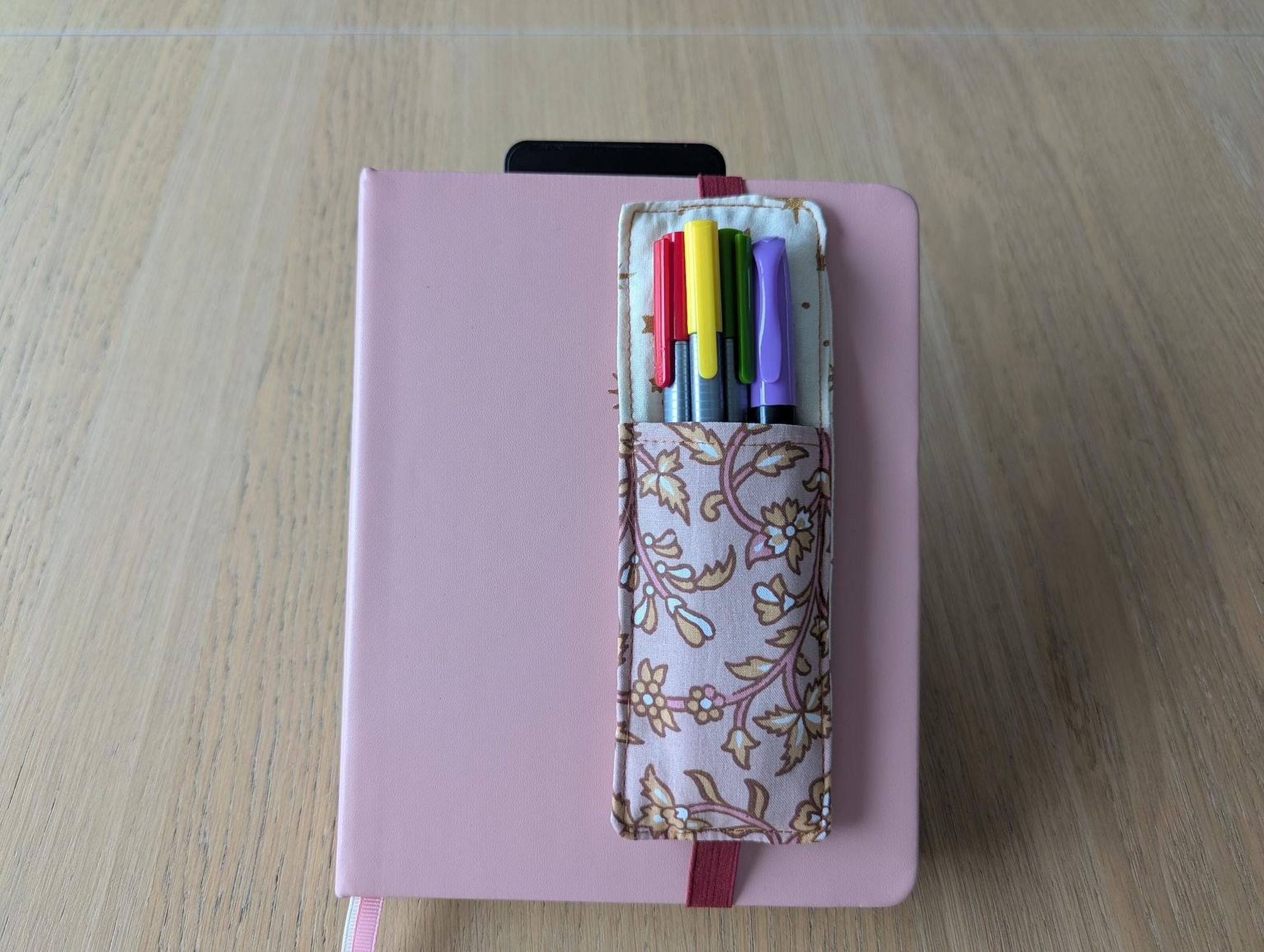 A pink notebook with a floral pattern and a pen holder containing various colored pens are placed on a wooden surface.