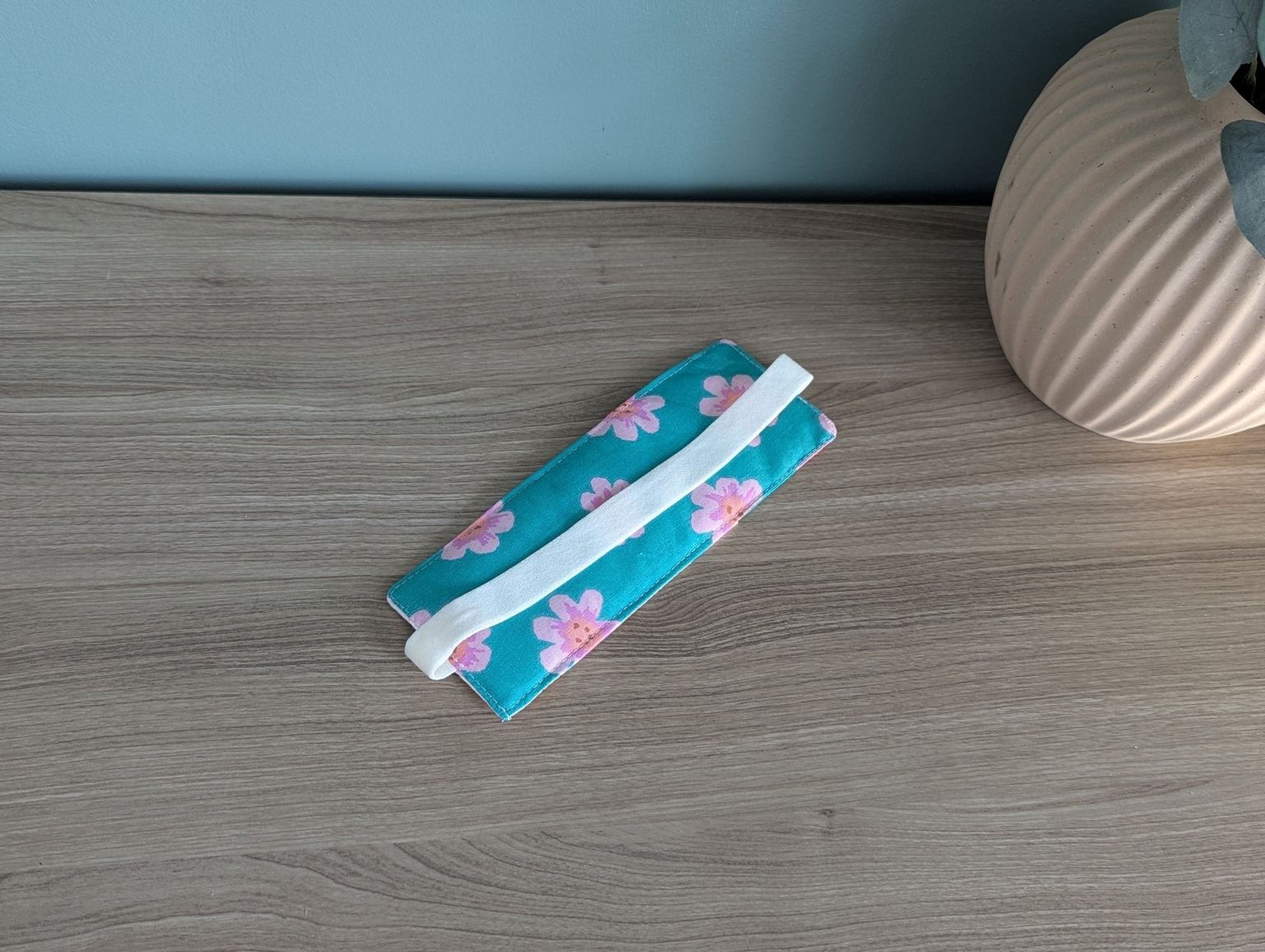 A blue and pink floral patterned fabric item, possibly a wallet or card holder, is placed on a wooden surface next to a vase.