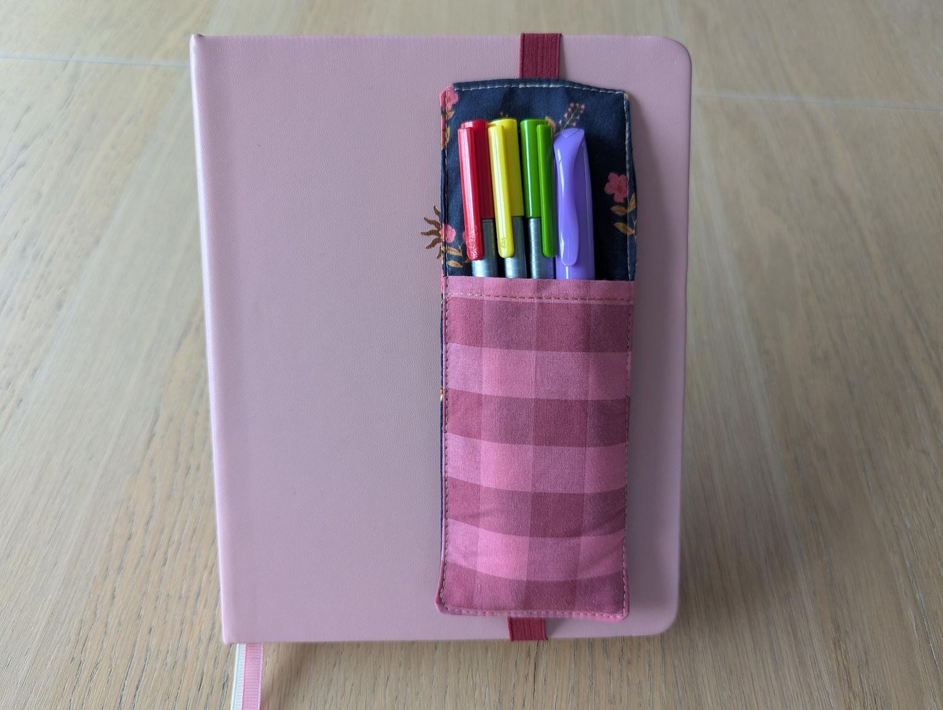 A pink notebook with a pocket attached to the cover, holding a set of colorful pens.
