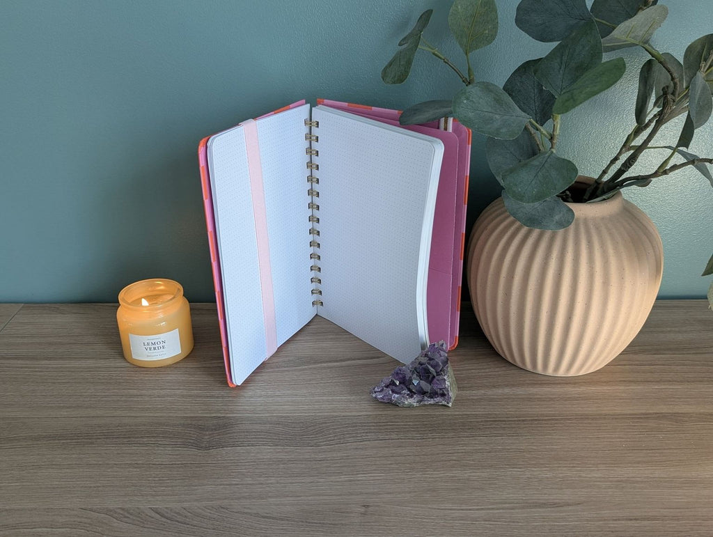 a wooden desk with a pink notebook, a candle, a plant, and a crystal on it.