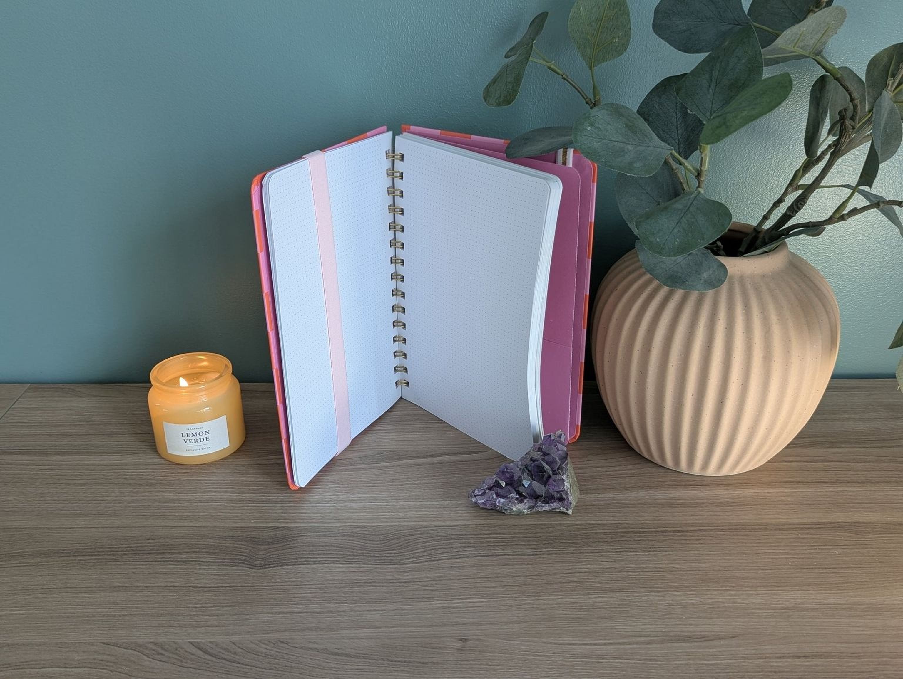 a wooden desk with a pink notebook, a candle, a plant, and a crystal on it.