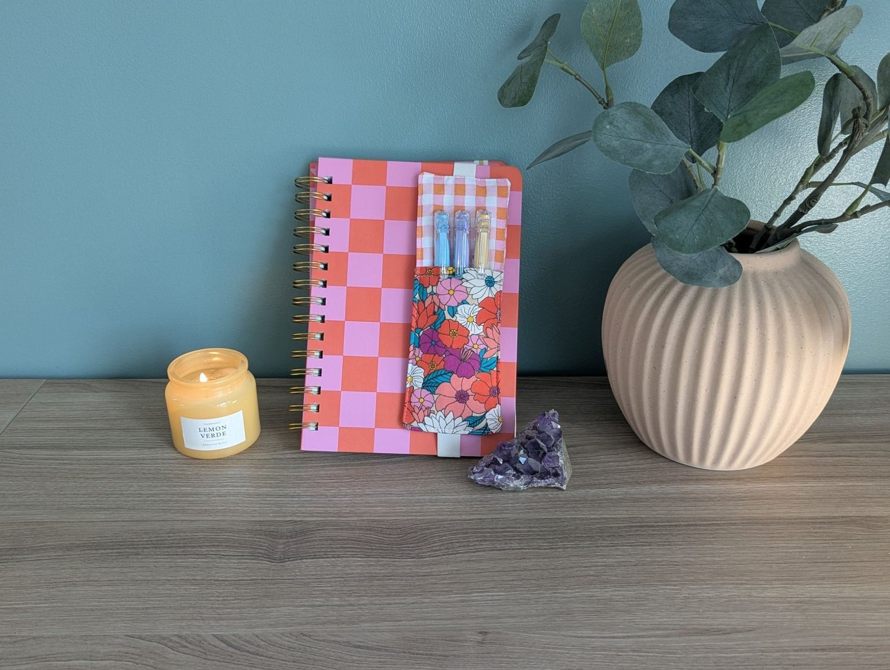 a pink and orange checkered notebook, a candle, a small crystal, and a beige vase with a plant on a wooden surface.