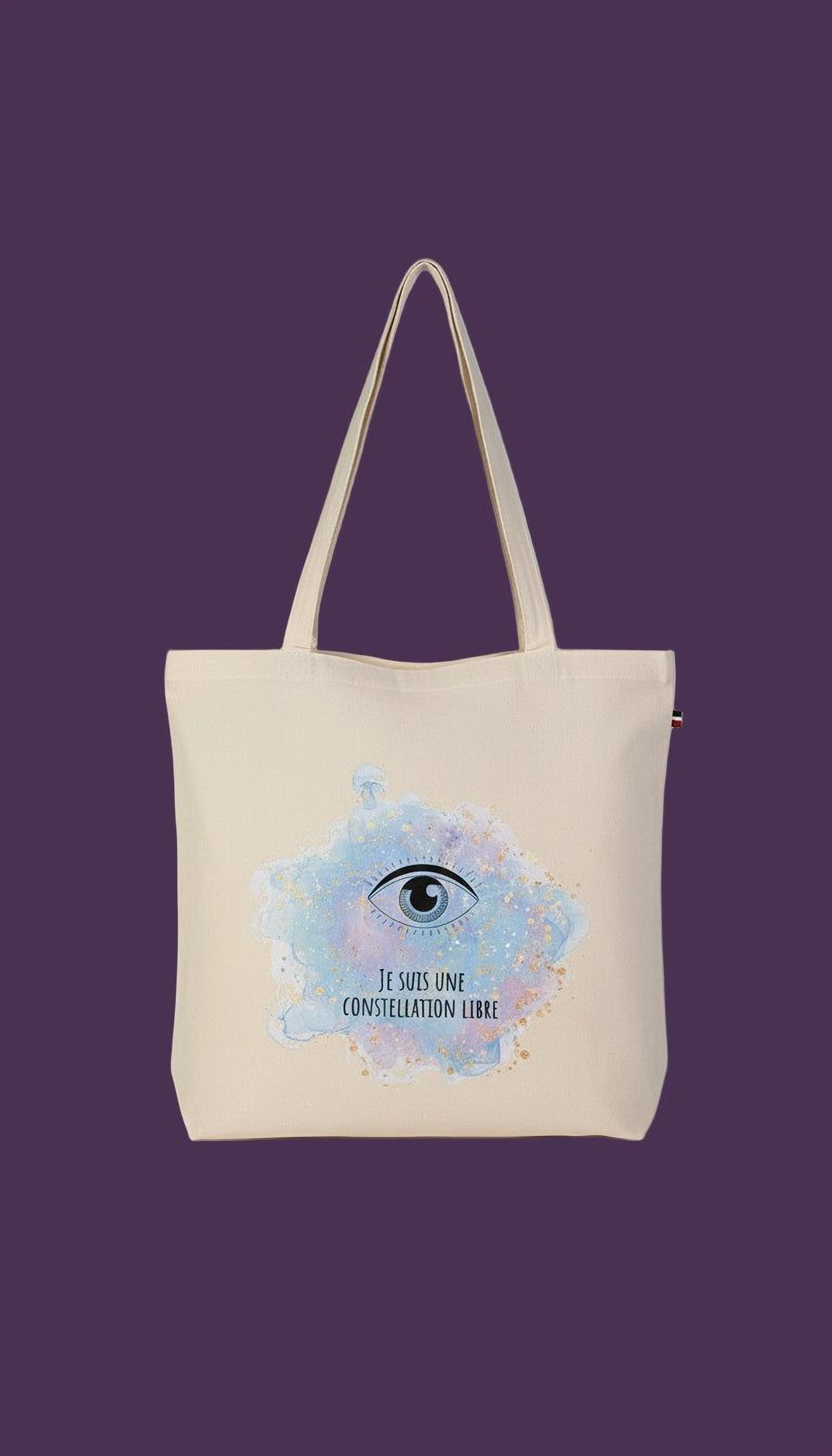 A beige tote bag with a watercolor illustration of an eye and the text "JE SUIS UNE CONSTELLATION LIBRE" printed on it.