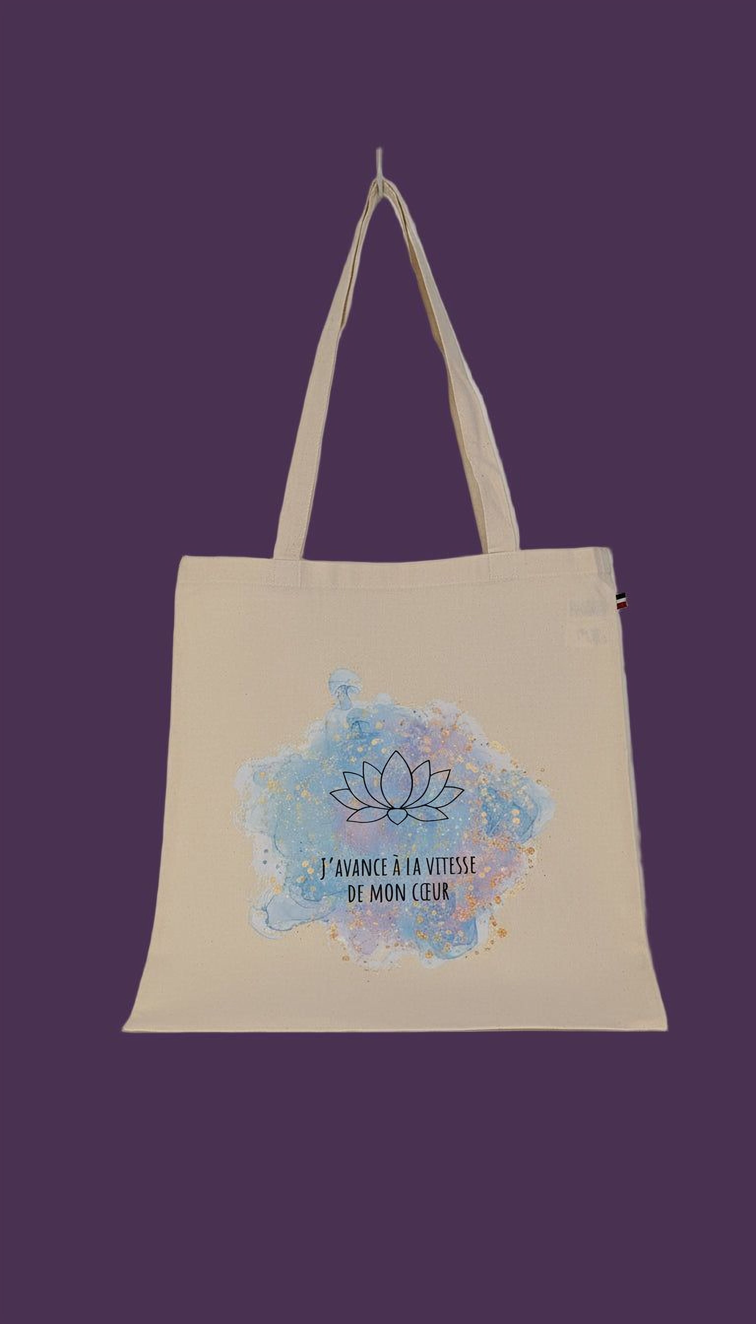A beige canvas tote bag with a blue and pink watercolor design featuring a lotus flower and the text "I'AVANCE A LA VITESSE DE MON CELUR" (I'm here to serve you).