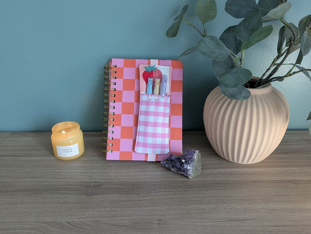 a wooden desk with a pink and orange checkered notebook, a candle, a small vase with a plant, and a crystal on it.