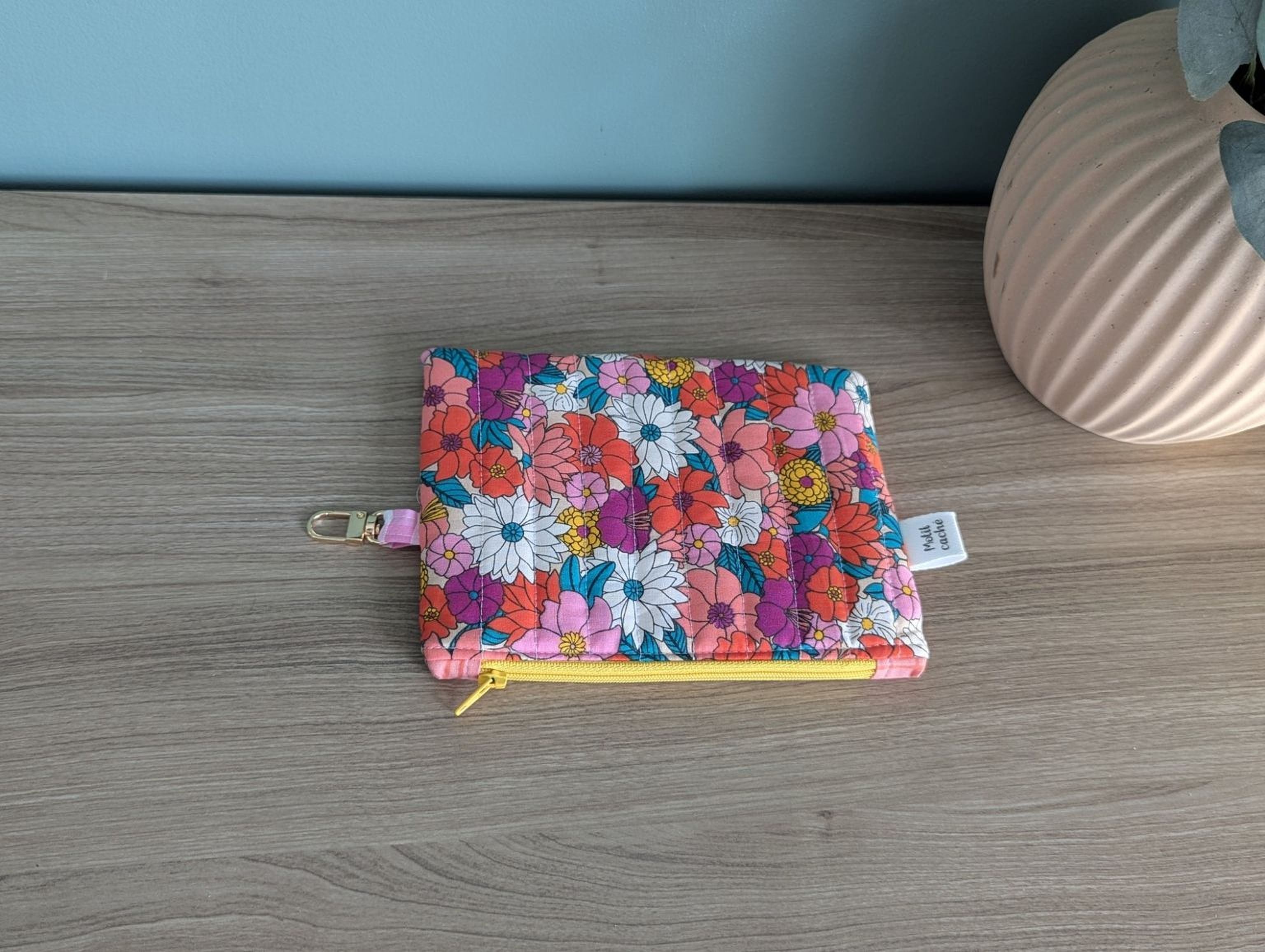 A colorful floral patterned pouch or wallet is placed on a wooden surface next to a vase.
