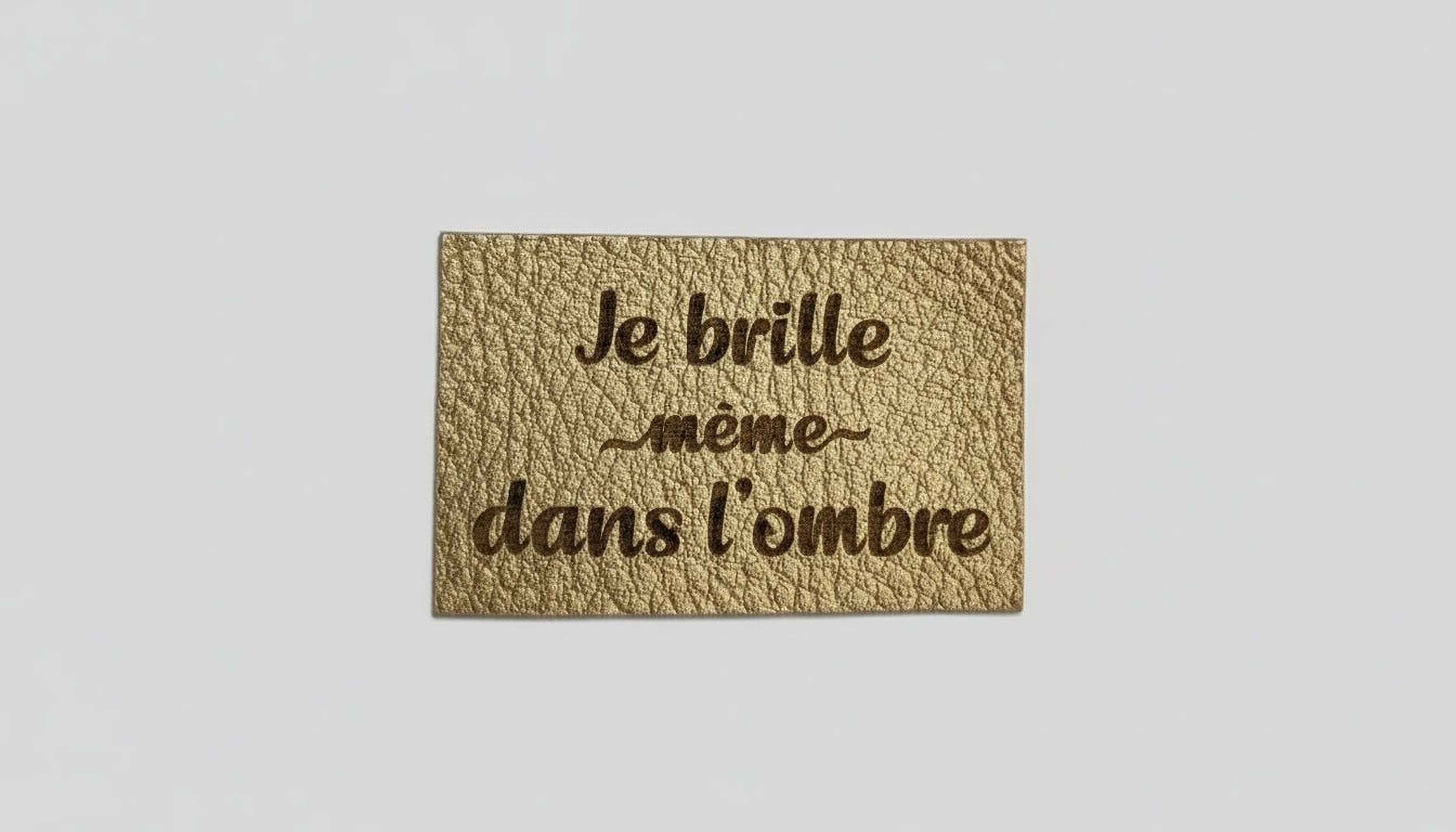 A gold-colored label with the text "Je brille même dans l'ombre" printed on it.
