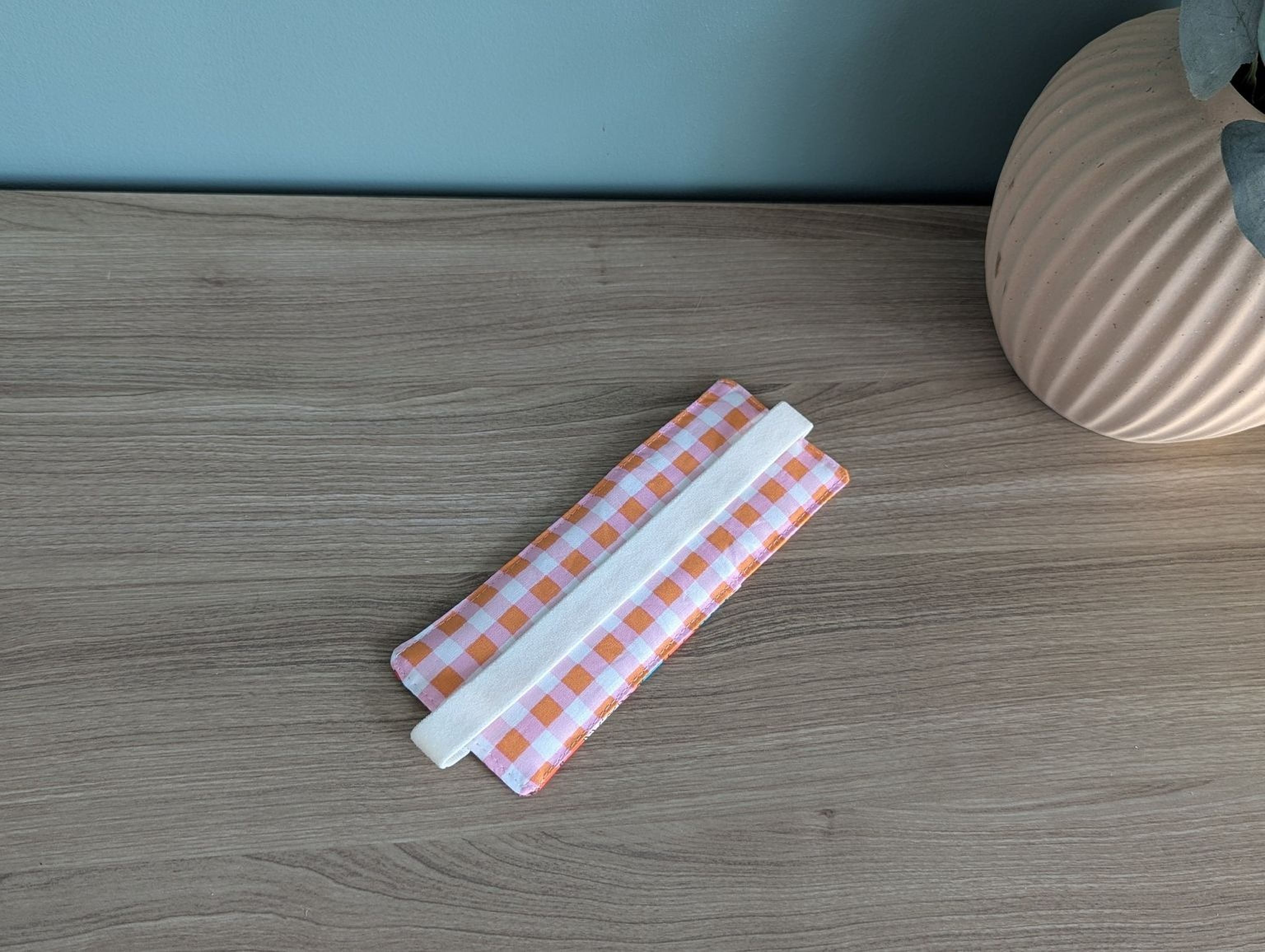 a wooden table with a white and orange checkered cloth or napkin placed on it. Next to the cloth, there is a small vase or container, possibly containing a decorative item or a small plant.