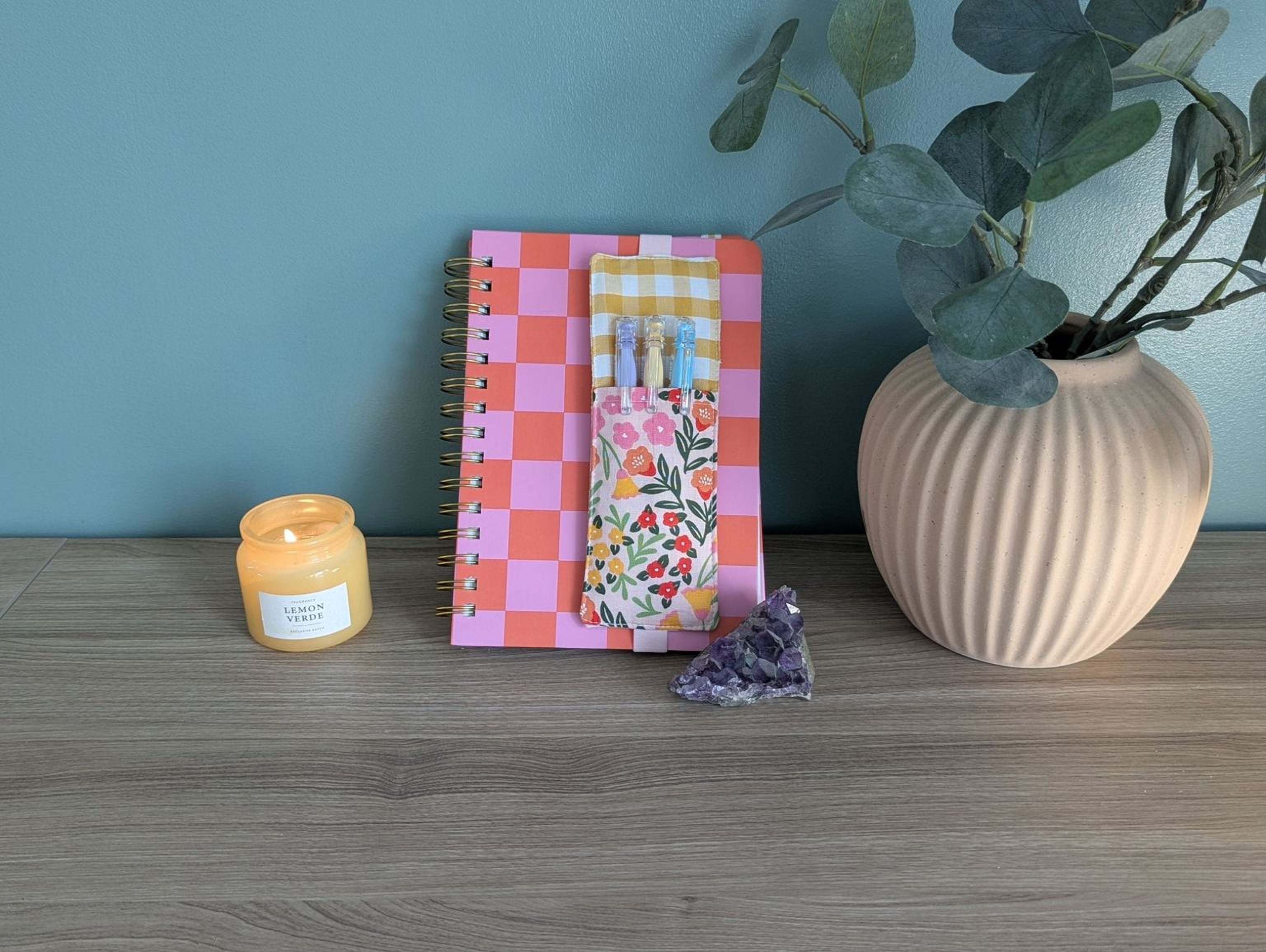 a wooden desk with a pink and orange checkered notebook, a candle, a small purple crystal, and a beige vase with a plant.