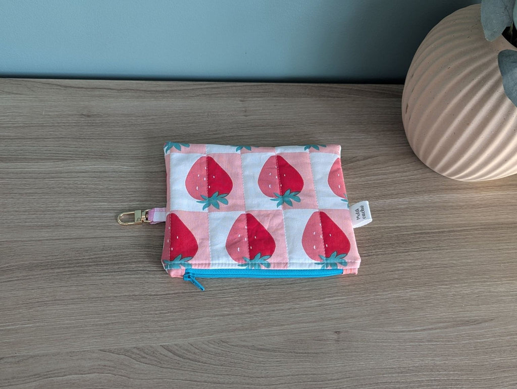 A small, colorful pouch with a strawberry pattern is placed on a wooden surface next to a vase.