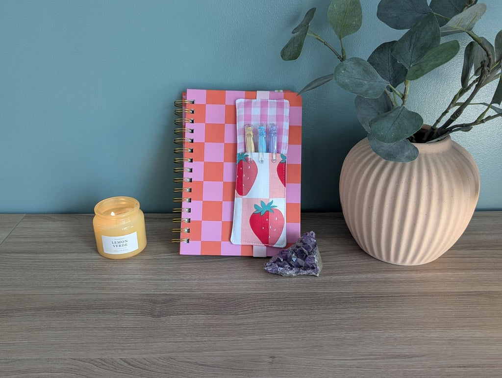 a wooden desk with a pink and orange checkered notebook, a candle, a small crystal, and a plant in a vase.